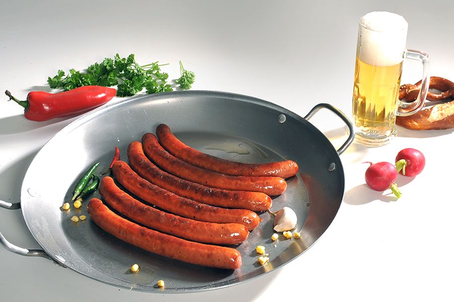 Fünf gebratene Würste in einer großen Pfanne mit Knoblauch, Chilischoten, Koriander, Maiskörnern, daneben ein Bierglas mit Schaum, eine Brezel und zwei Radieschen auf weißem Untergrund.